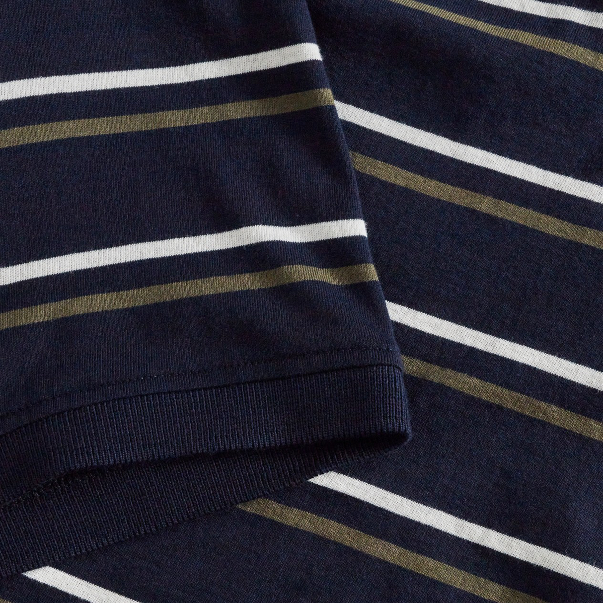 Close-up of a navy blue fabric with horizontal white and olive green stripes, characteristic of the Canterbury Yarn Dye Stripe Polo. The image features a sleeve overlapping the main body of the garment, evoking classic Canterbury rugby polo styling.