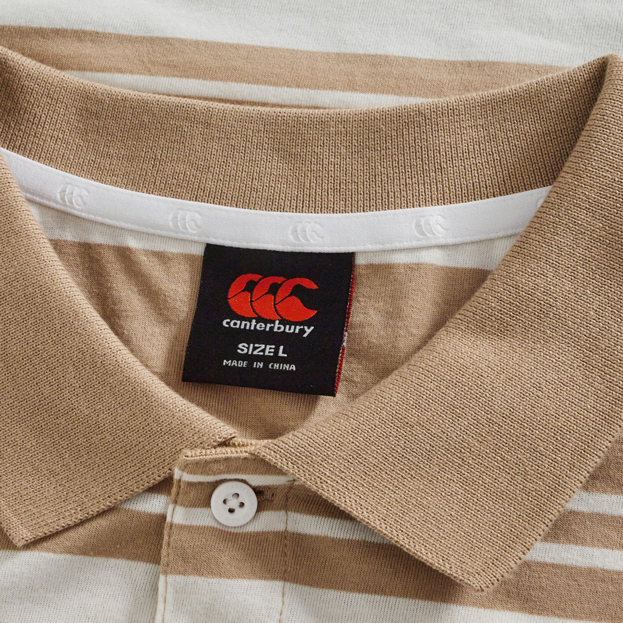 Close-up of the Fundamental Engineered Stripe Polo by Canterbury, featuring beige and white stripes, a classic collar, button detail, and a visible black Canterbury brand tag indicating size L and made in China.