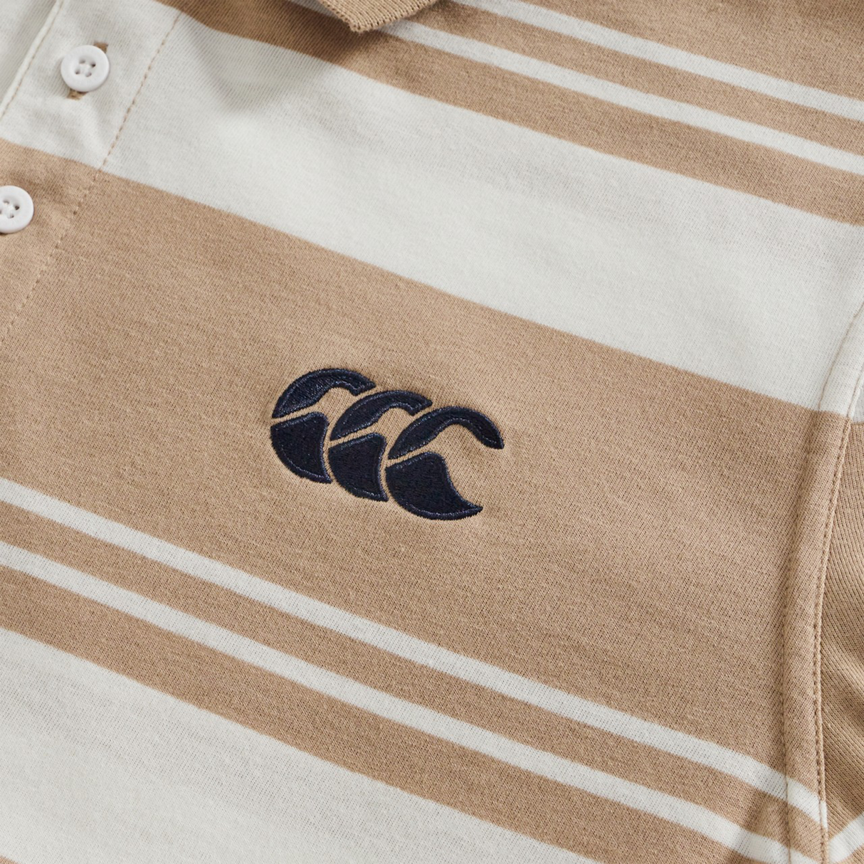 Close-up of the Fundamental Engineered Stripe Polo by Canterbury in beige and white, featuring three white buttons and a black embroidered logo with three overlapping "C" shapes.