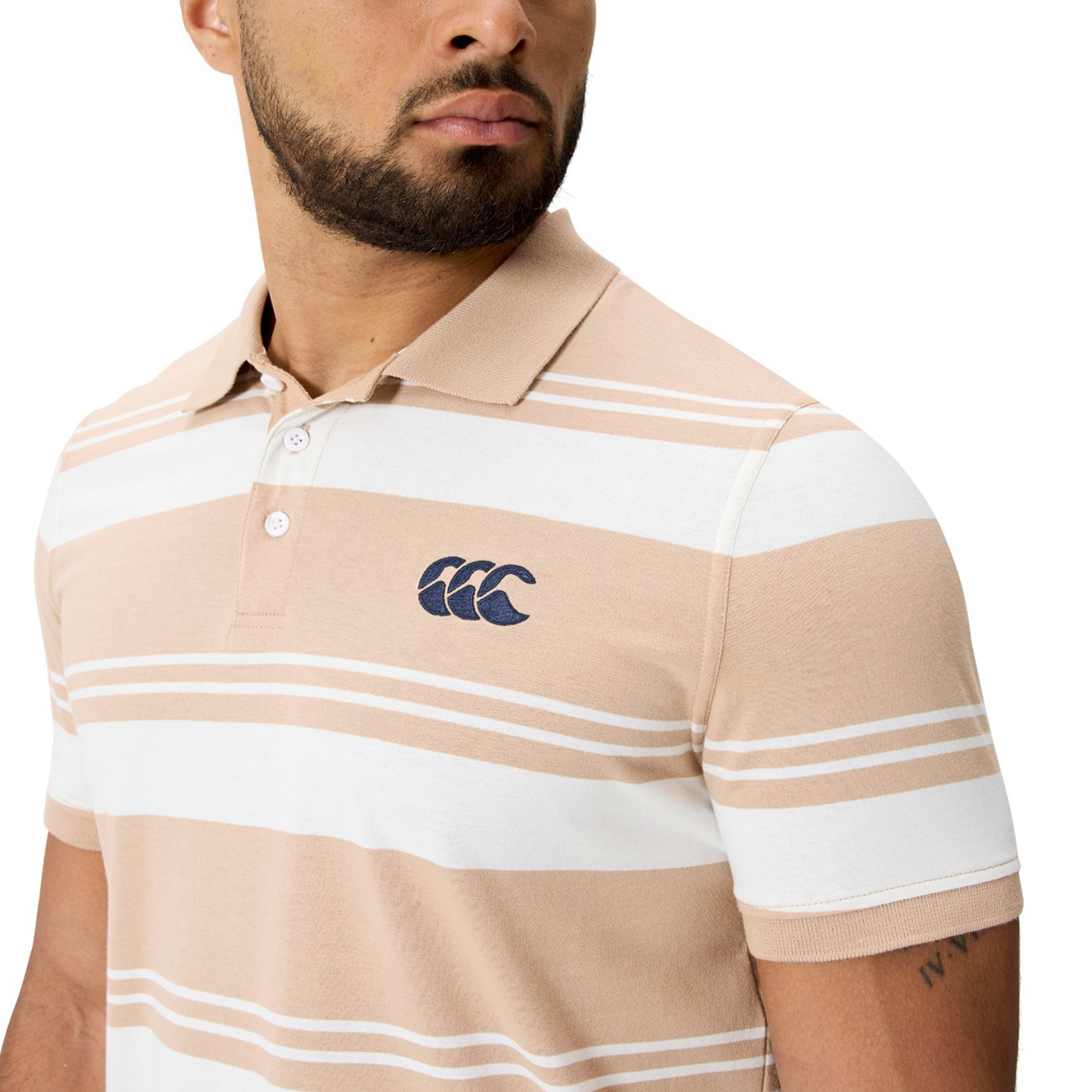 A man wearing the Canterbury Fundamental Engineered Stripe Polo in beige and white with a blue embroidered logo on the chest, looking to the side.