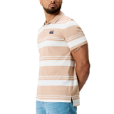 A man stands with his arms behind his back, wearing the Fundamental Engineered Stripe Polo by Canterbury in beige and white with a chest logo, paired with light blue jeans.