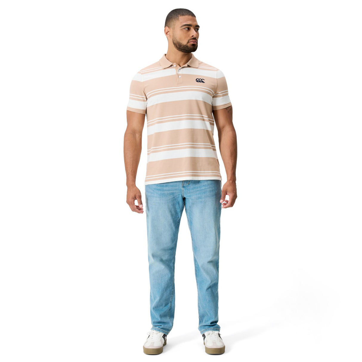A man faces forward wearing the Fundamental Engineered Stripe Polo by Canterbury in beige and white, paired with light blue jeans and white sneakers, set against a plain white background.
