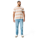 A man faces forward wearing the Fundamental Engineered Stripe Polo by Canterbury in beige and white, paired with light blue jeans and white sneakers, set against a plain white background.