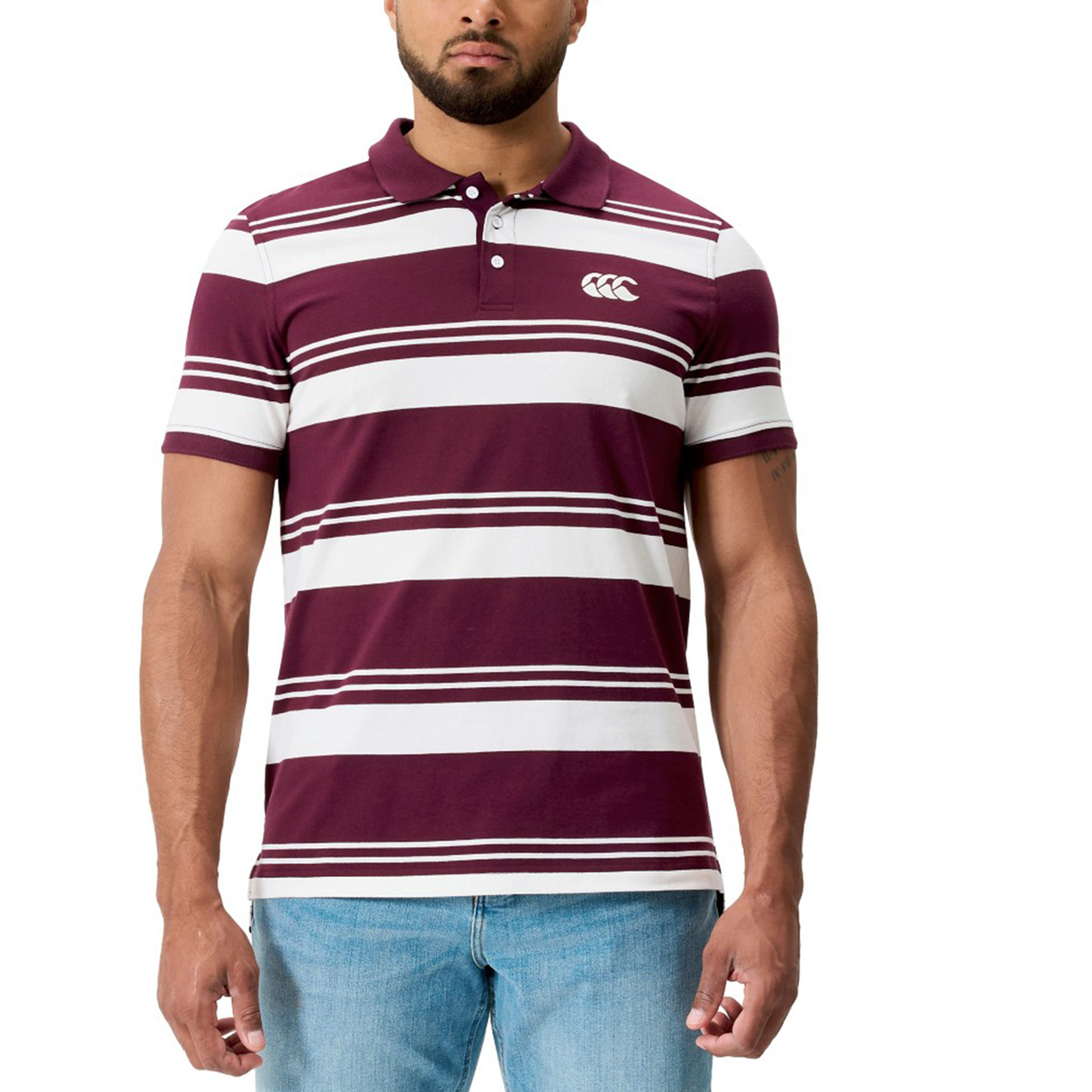 A man wears the Fundamental Engineered Stripe Polo by Canterbury, featuring maroon and white rugby-inspired stripes with a chest logo, paired with light blue jeans.