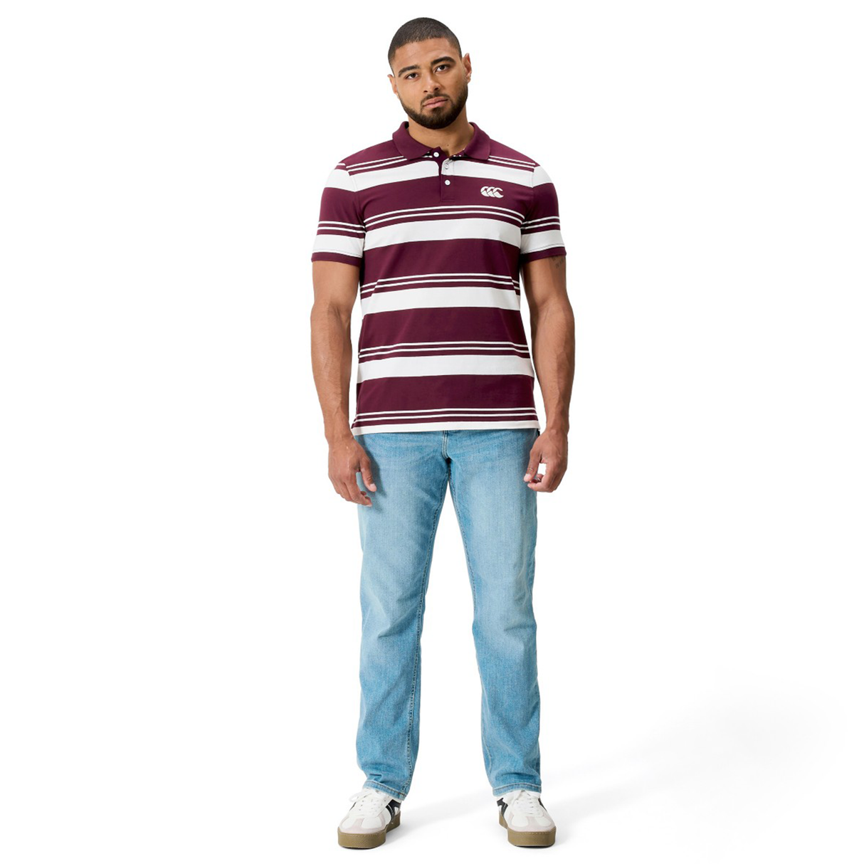 A man stands against a white background wearing the Canterbury Fundamental Engineered Stripe Polo inspired by rugby heritage, paired with light blue jeans and white sneakers.
