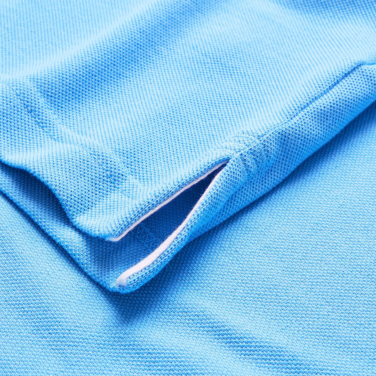 Here’s a revised version using the product data: Close-up of a light blue textured fabric from the Canterbury by Canterbury Solid Dye Polo, highlighting a small side slit and precise stitching along the seam.