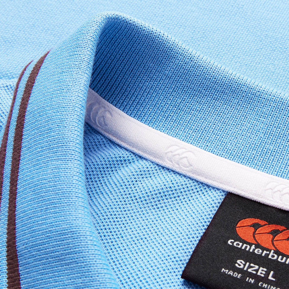 Close-up of the light blue Canterbury Solid Dye Polo, showcasing brown and white stripes on the collar and a visible black Canterbury size label.
