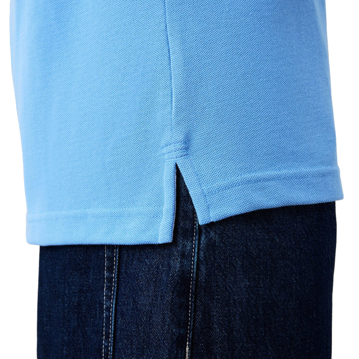Close-up of the hem of the Canterbury Solid Dye Polo by Canterbury in light blue, featuring a small side slit, worn over dark blue denim jeans.