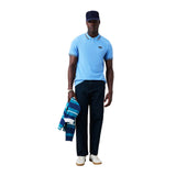 A man dressed in a blue Canterbury Solid Dye Polo by Canterbury, paired with dark jeans, white sneakers, and a navy cap, stands against a white background while holding a striped jacket.