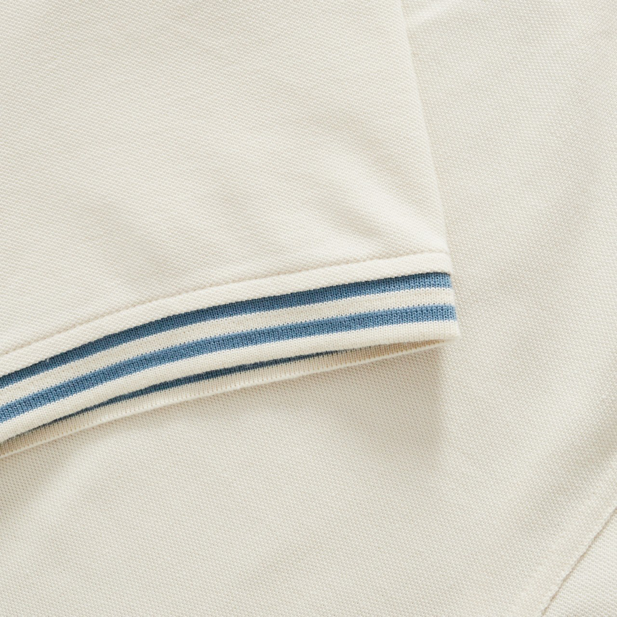 Close-up of the Canterbury Fundamental Solid Dye Polo in beige, featuring short sleeves and blue-white striped trim, highlighting its rugby-inspired design, displayed flat.