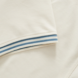 Close-up of the Canterbury Fundamental Solid Dye Polo in beige, featuring short sleeves and blue-white striped trim, highlighting its rugby-inspired design, displayed flat.