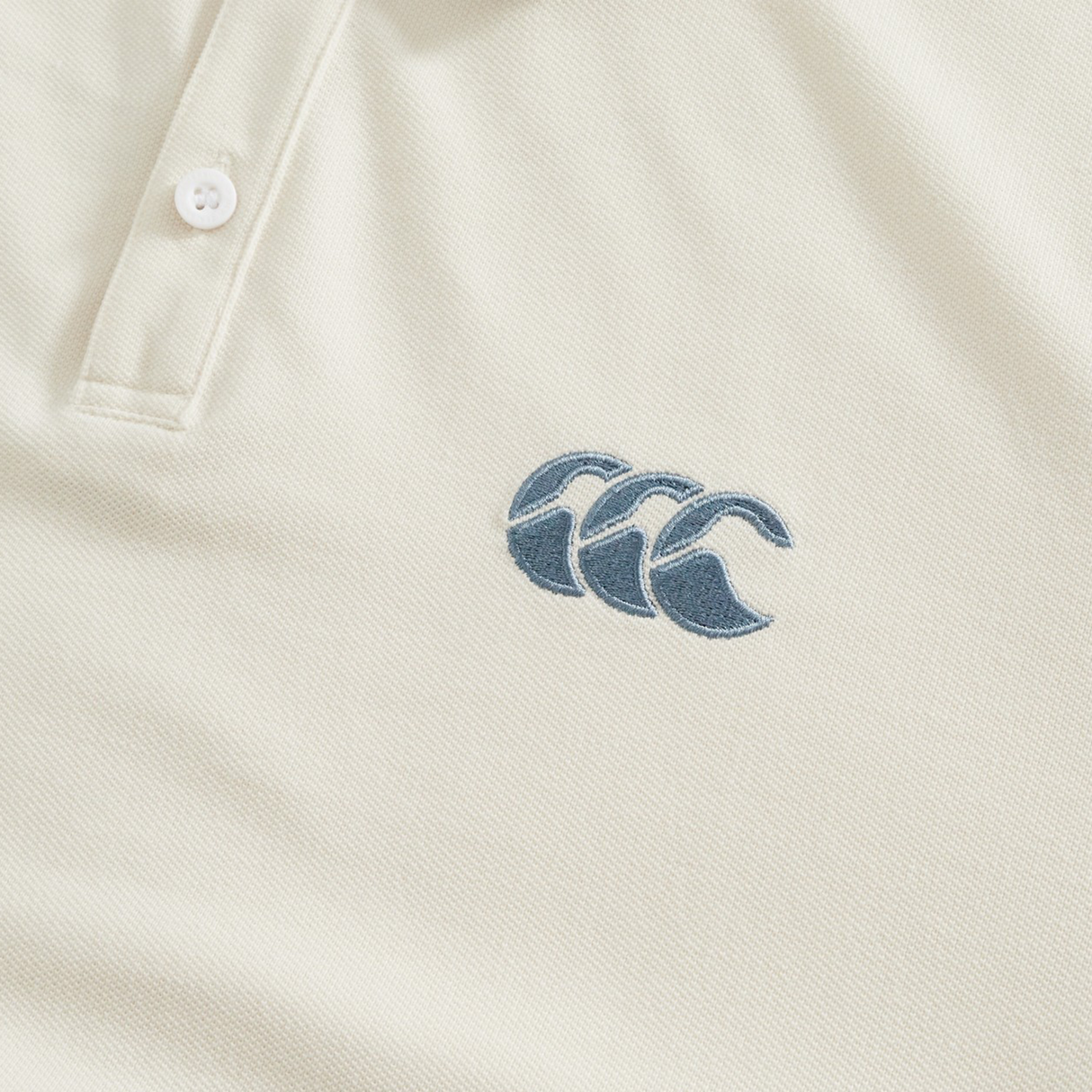 Close-up of the cream-colored Fundamental Solid Dye Polo by Canterbury, featuring a stitched blue logo with three overlapping waves on the chest, showcasing its subtle polo and rugby-inspired design.