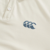 Close-up of the cream-colored Fundamental Solid Dye Polo by Canterbury, featuring a stitched blue logo with three overlapping waves on the chest, showcasing its subtle polo and rugby-inspired design.