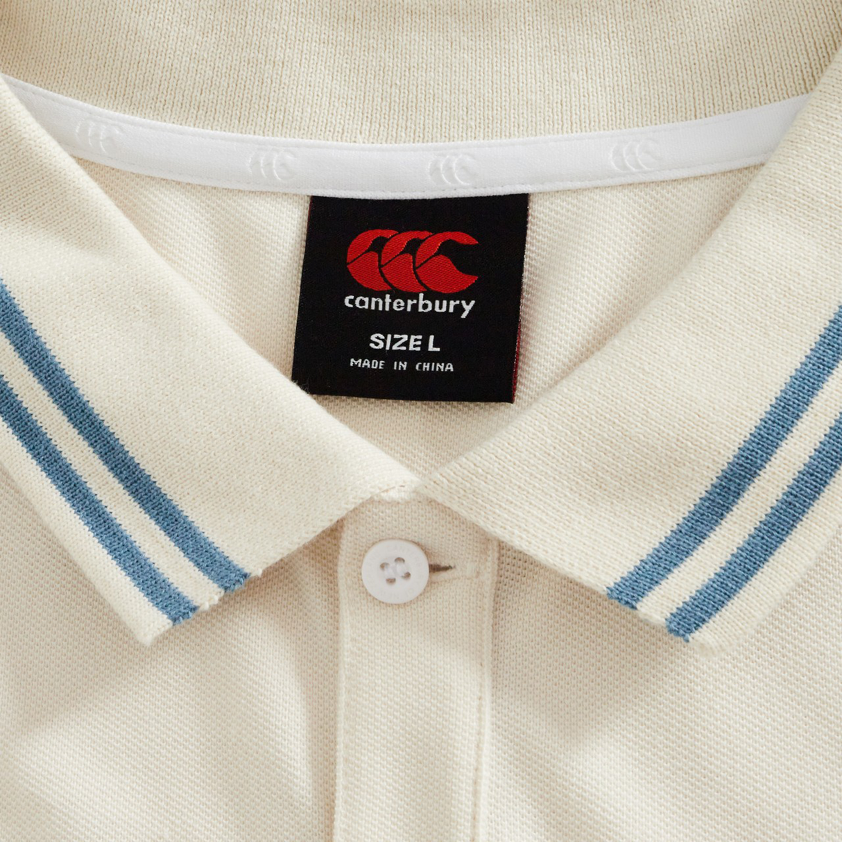 Close-up of the Canterbury Fundamental Solid Dye Polo: cream color, blue-striped collar, white button, and black brand label showing size L, made in China.