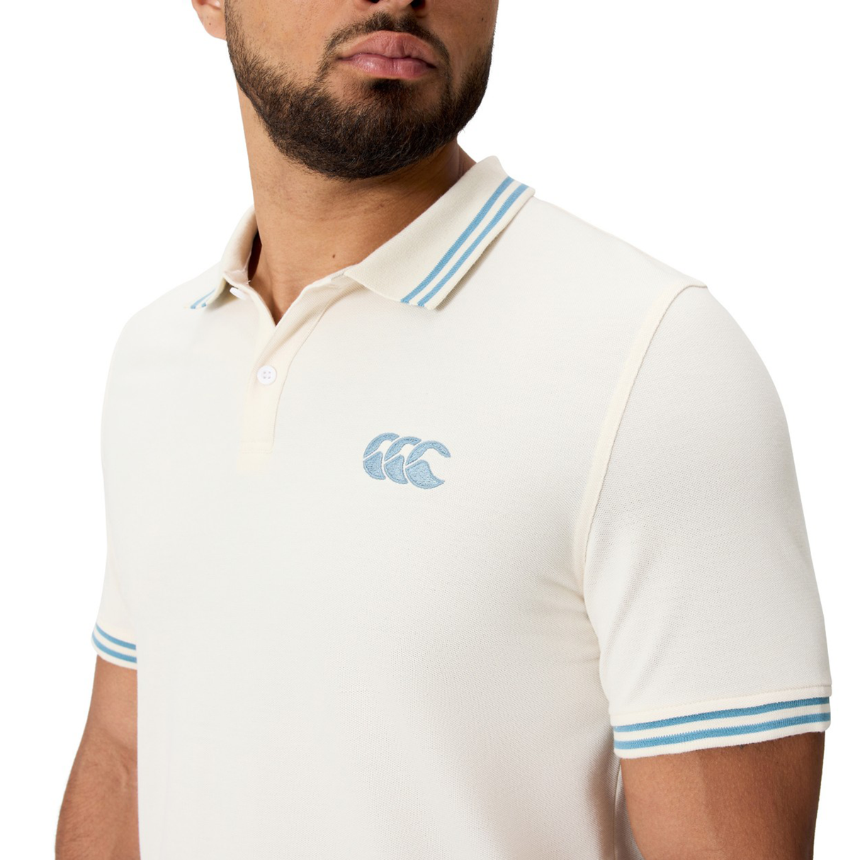 A man wearing Canterbury’s Fundamental Solid Dye Polo in cream, featuring a blue and white striped collar and cuffs, plus a blue embroidered chest logo—capturing classic rugby-inspired style.