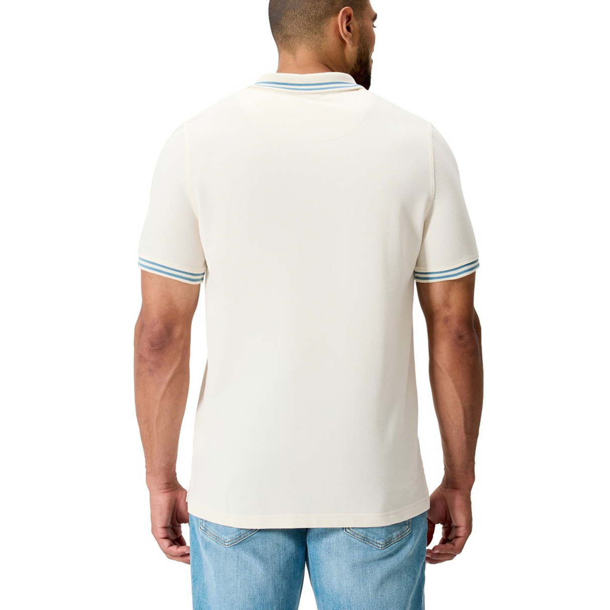 A man is seen from the back in light blue jeans and a cream Fundamental Solid Dye Polo by Canterbury, featuring green-striped cuffs; the rugby-inspired design adds a sporty edge.