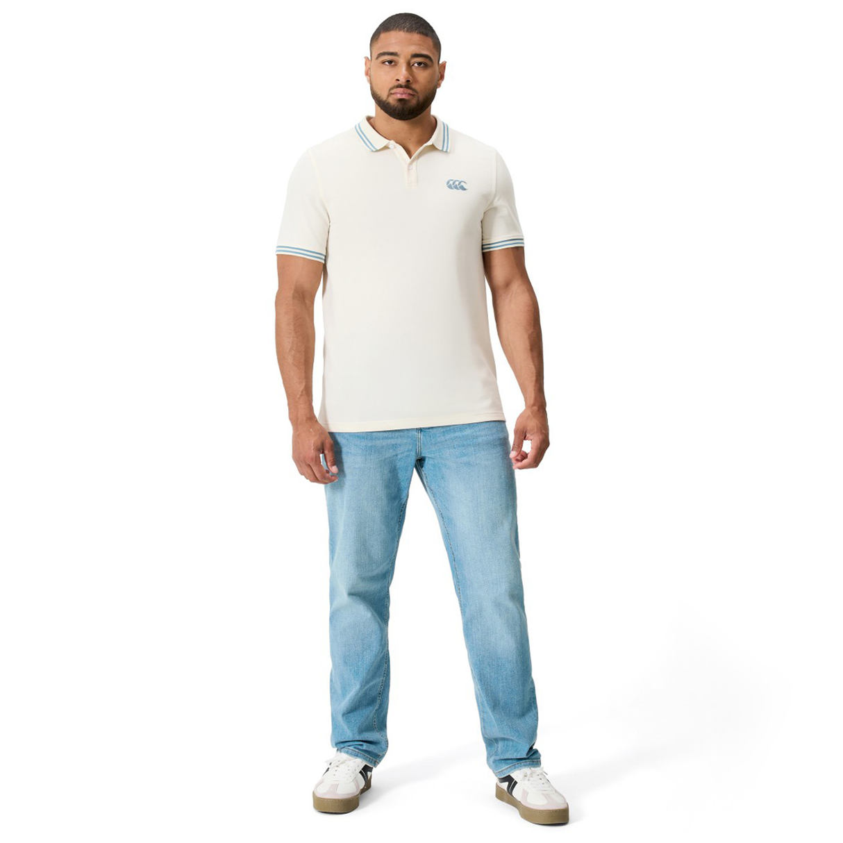 A man stands facing forward against a white background, wearing the Canterbury Fundamental Solid Dye Polo in cream, paired with light blue jeans and white sneakers.