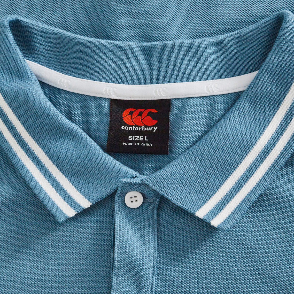 Close-up of the Canterbury Fundamental Solid Dye Polo in blue, featuring a white-striped collar, white button, and black size L label—highlighting its classic rugby-inspired style.