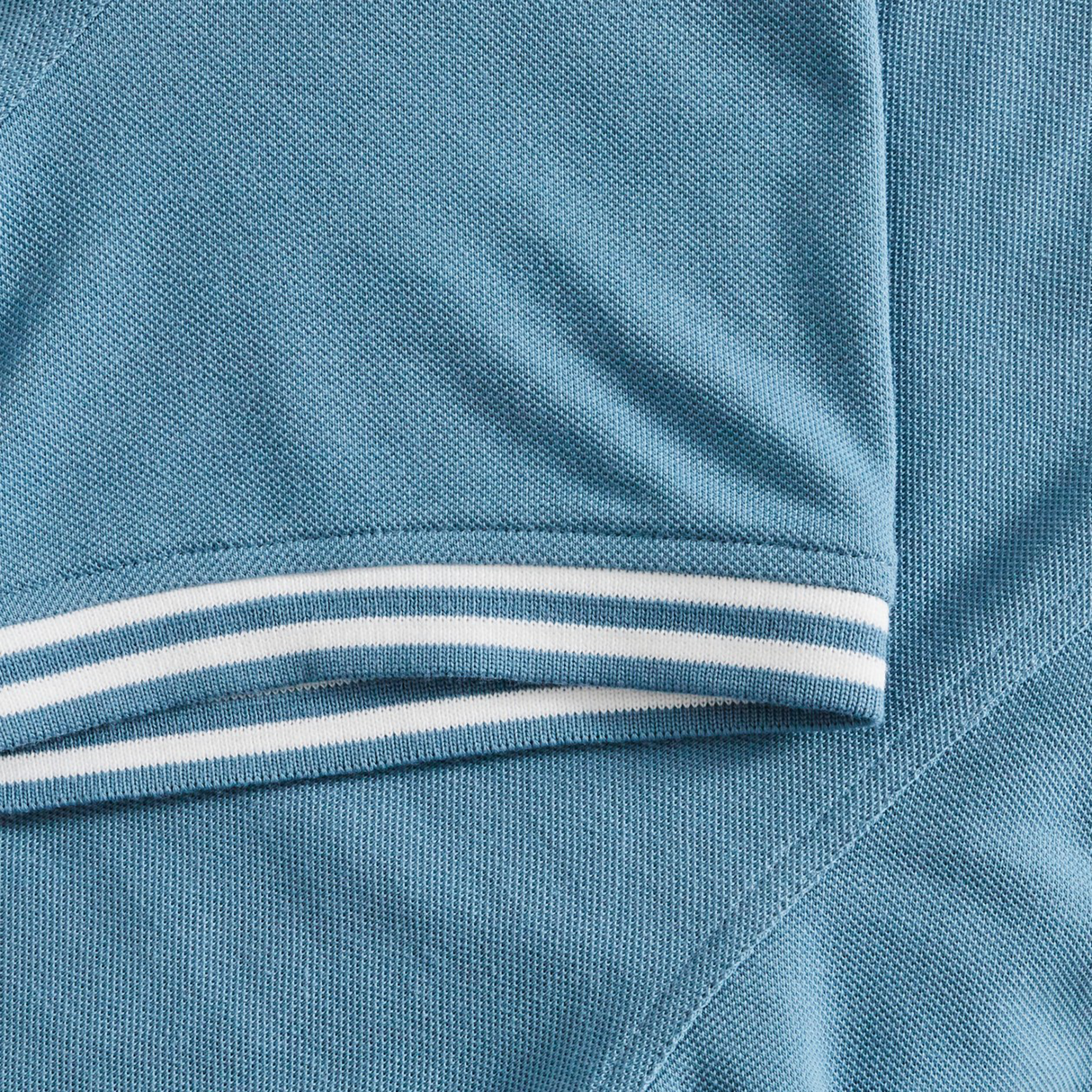 Close-up of the Fundamental Solid Dye Polo by Canterbury, a blue short-sleeve shirt featuring white and blue striped trim at the cuff for a subtle rugby-inspired look.