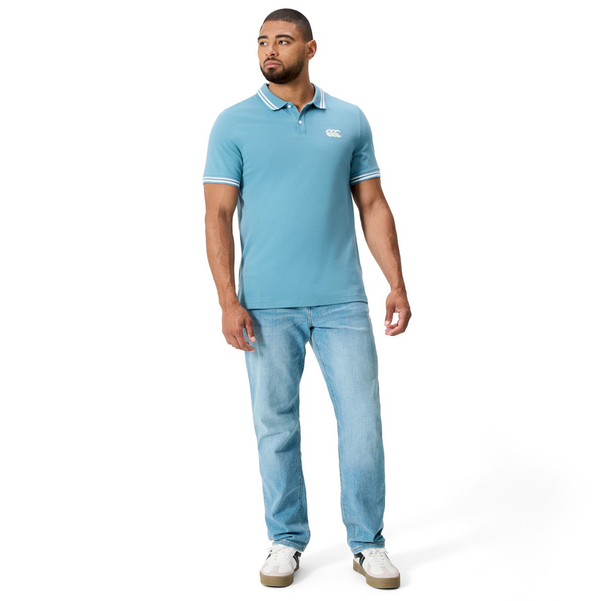 A man faces slightly to the side, wearing the Canterbury Fundamental Solid Dye Polo in light blue, paired with light blue jeans and white sneakers, set against a plain white background.