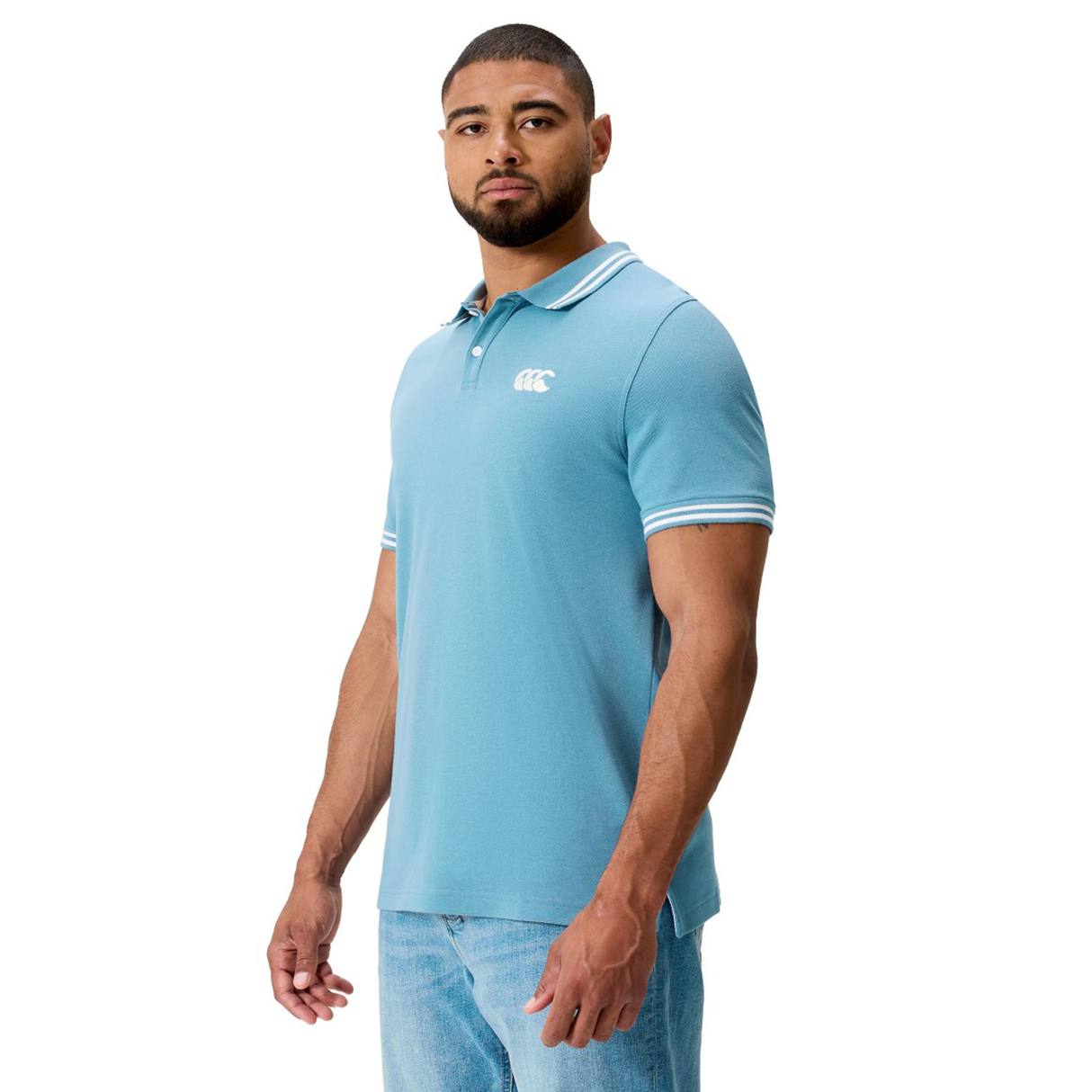 A man wears the Canterbury Fundamental Solid Dye Polo in light blue with white trim and blue jeans, standing slightly turned against a white background.