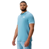 A man wears the Canterbury Fundamental Solid Dye Polo in light blue with white trim and blue jeans, standing slightly turned against a white background.