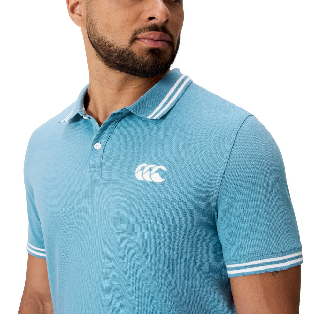 A man wearing Canterbury’s Fundamental Solid Dye Polo—a light blue, short-sleeve shirt with white trim and a white chest logo.