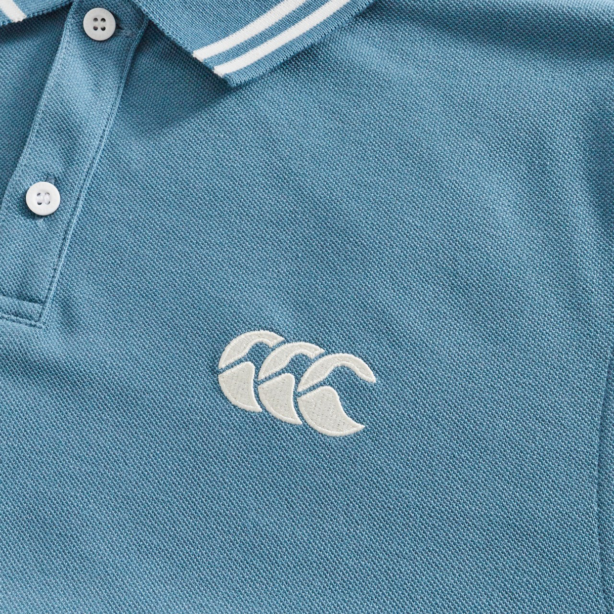 Close-up of the Fundamental Solid Dye Polo by Canterbury, featuring white buttons, a striped collar, and an embroidered chest logo—classic rugby-inspired style from Canterbury.