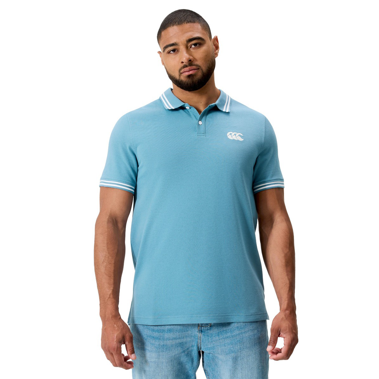 A man wears the Fundamental Solid Dye Polo by Canterbury in light blue with white trim and light blue jeans, standing against a plain white background.