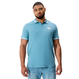 A man wears the Fundamental Solid Dye Polo by Canterbury in light blue with white trim and light blue jeans, standing against a plain white background.