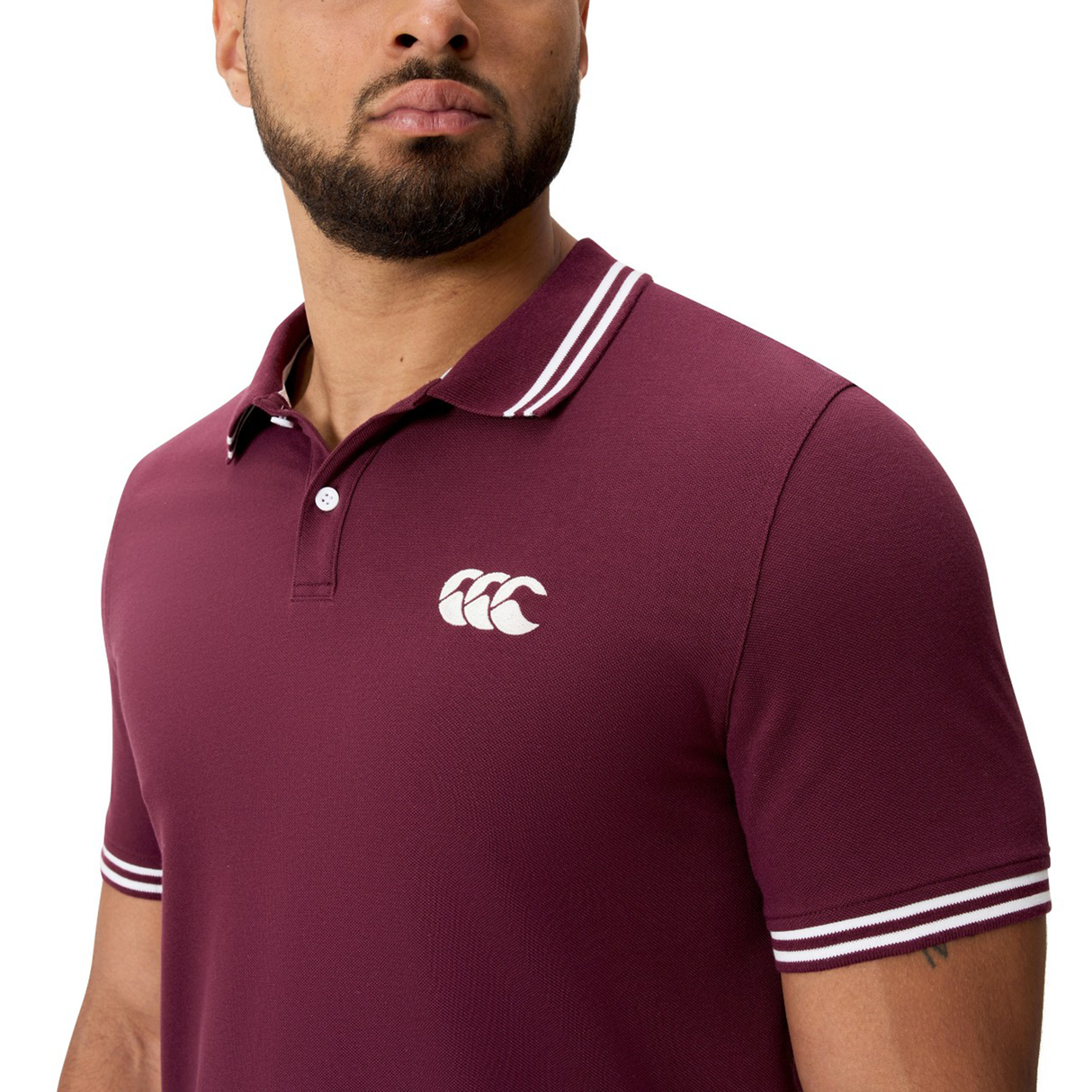 A man wears the Canterbury Fundamental Solid Dye Polo, a maroon rugby-inspired shirt with white stripes on the collar and sleeves, and a white chest logo.