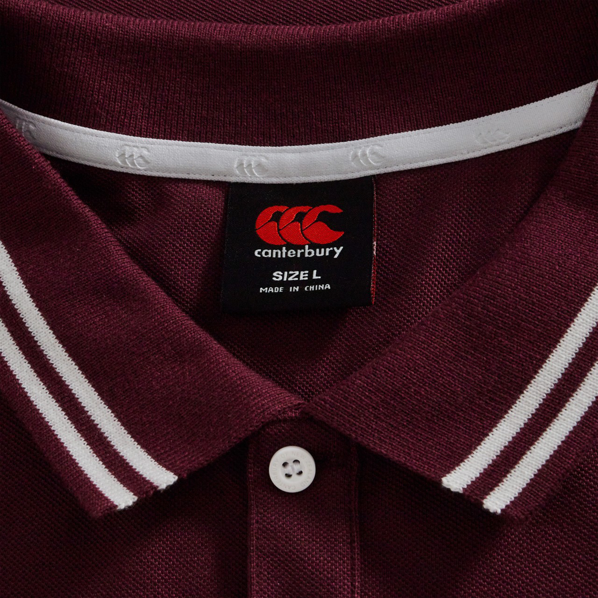 Close-up of the Canterbury Fundamental Solid Dye Polo in maroon, featuring white-striped collar, rugby-inspired design, and a visible tag showing size L and made in China.