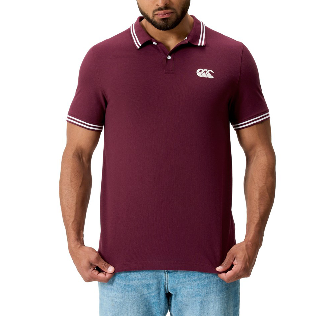 A man wearing Canterbury's Fundamental Solid Dye Polo in maroon with white trim, a rugby-inspired design, white chest logo, paired with light blue jeans.
