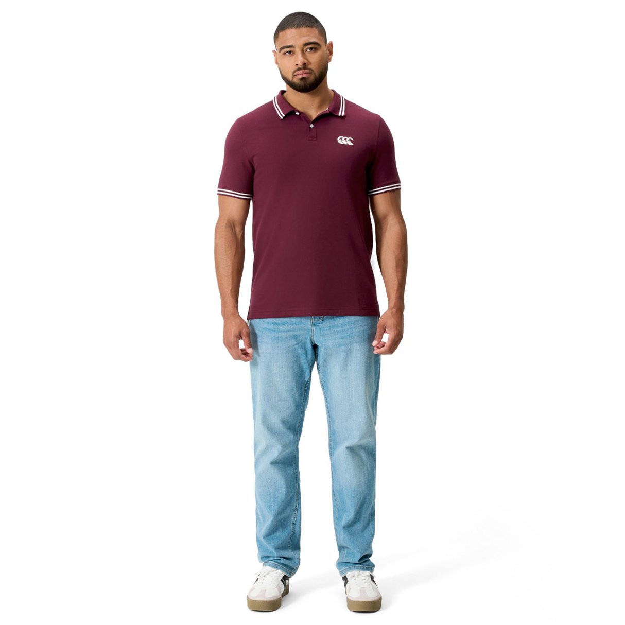 A man stands facing forward against a white background, wearing Canterbury's Fundamental Solid Dye Polo in maroon, paired with light blue jeans and white sneakers.