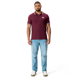 A man stands facing forward against a white background, wearing Canterbury's Fundamental Solid Dye Polo in maroon, paired with light blue jeans and white sneakers.