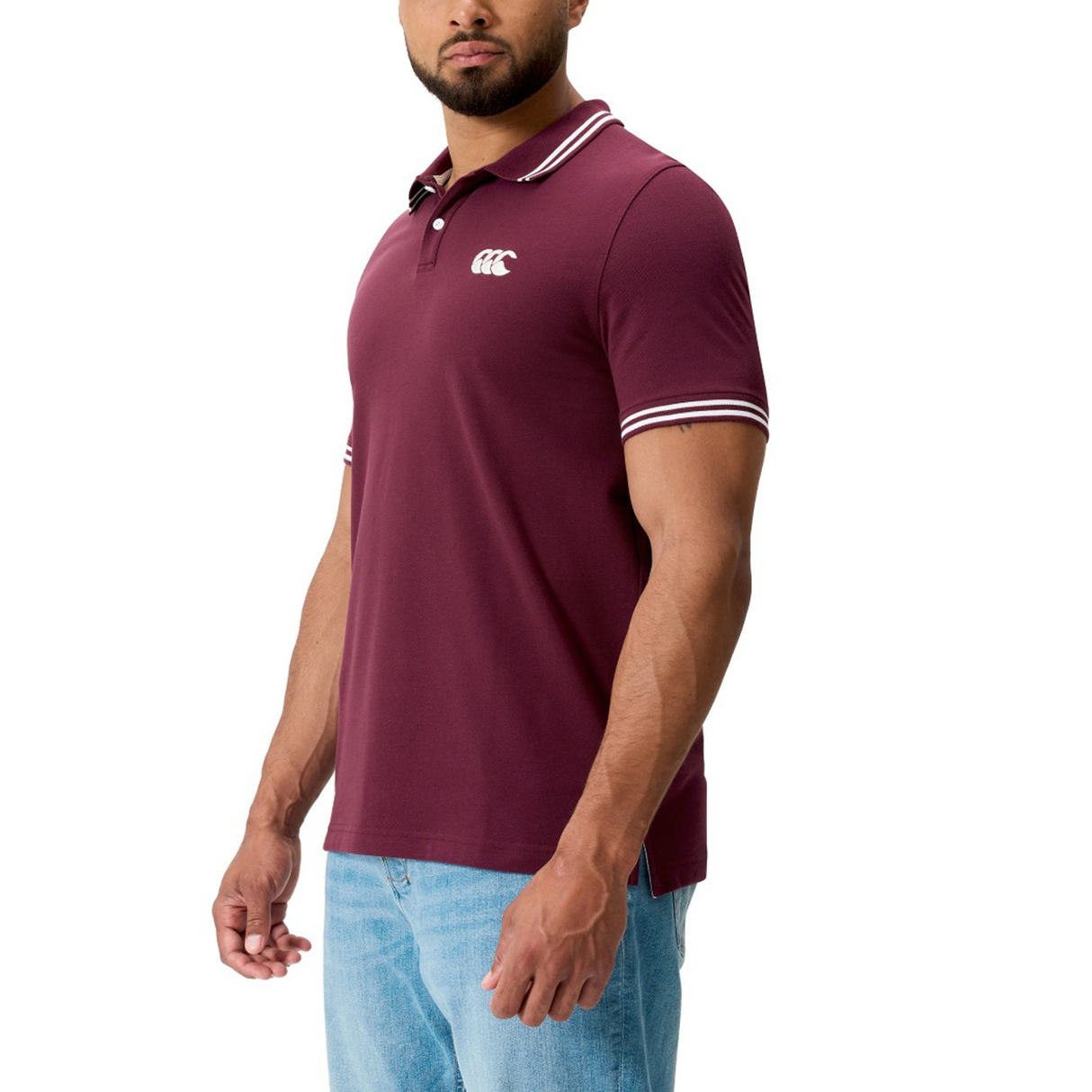 A man in the Canterbury Fundamental Solid Dye Polo, featuring white striped details and paired with light blue jeans, stands against a white background, showcasing this rugby-inspired Canterbury design.