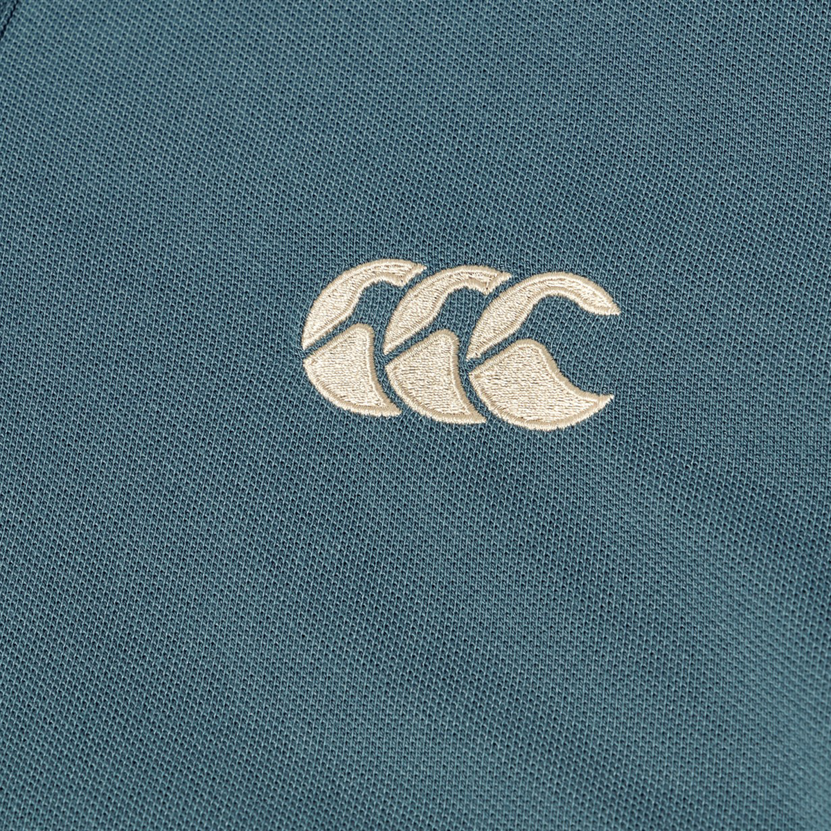 Close-up of a teal fabric from the Canterbury Solid Dye Polo, featuring an embroidered white Canterbury logo with three overlapping curved shapes, capturing the athletic style of this classic polo.
