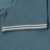 Close-up of the Canterbury Solid Dye Polo by Canterbury, featuring short sleeves in a rich teal color and a beige and white striped cuff, highlighting its rugby-inspired athletic style with detailed fabric texture and precise stitching.