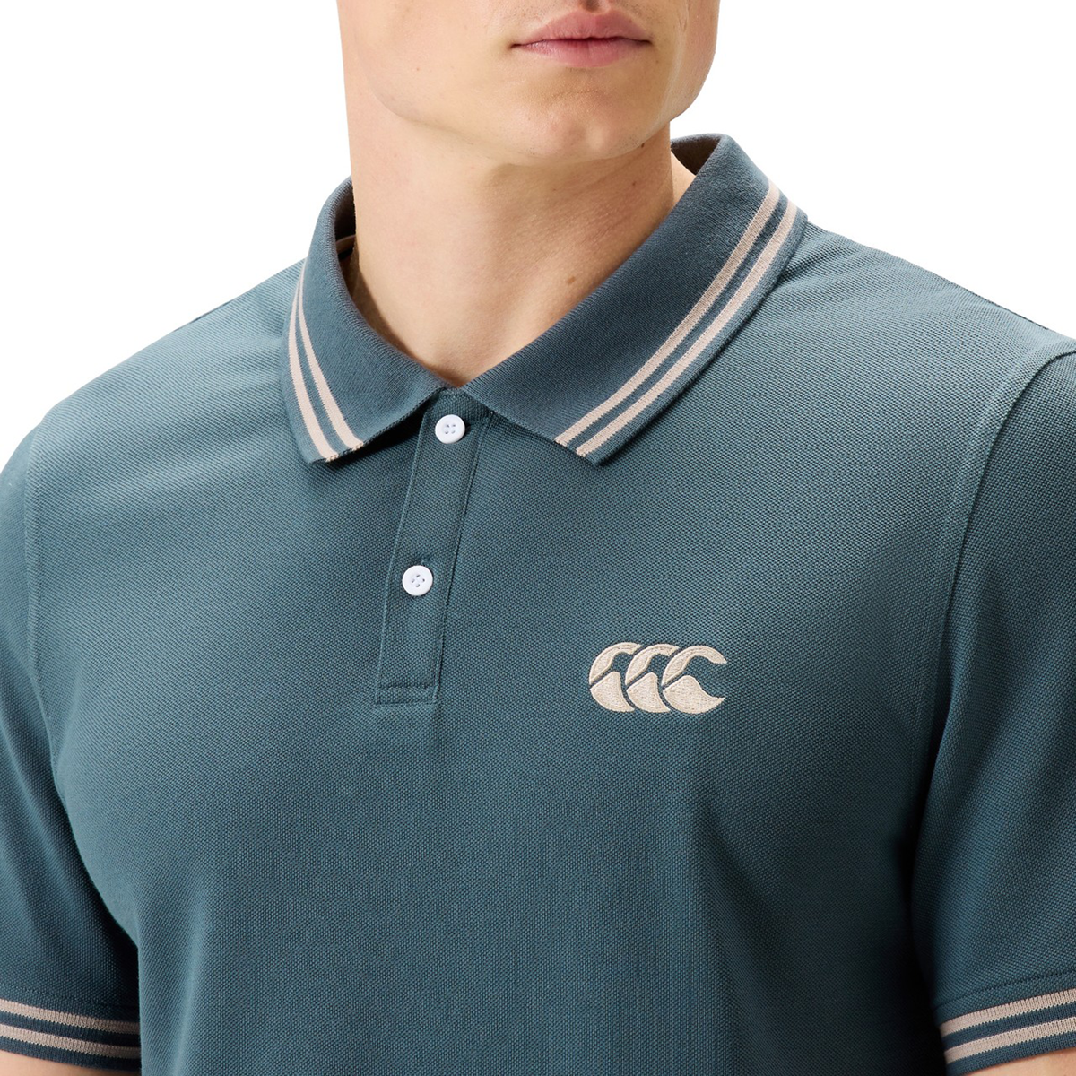 A person wearing the Canterbury Solid Dye Polo in teal, featuring a white Canterbury logo and a striped collar, visible from shoulders to chest, exemplifying a rugby-inspired athletic style.