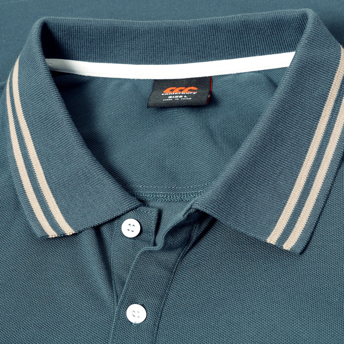 Close-up of the Canterbury Solid Dye Polo, a teal rugby-inspired shirt by Canterbury, featuring beige stripes on the collar, two white buttons, and a visible black brand tag under the collar.