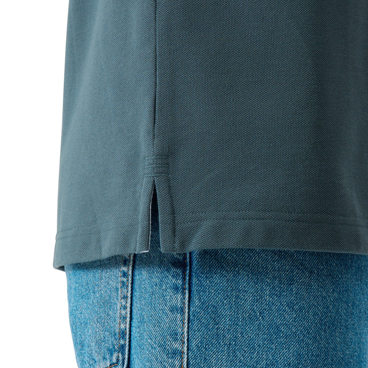 Close-up of the Canterbury Solid Dye Polo by Canterbury in teal, featuring a rugby-inspired design and a small side slit at the hem, styled with blue denim jeans.