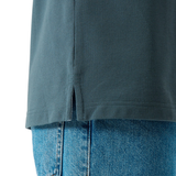 Close-up of the Canterbury Solid Dye Polo by Canterbury in teal, featuring a rugby-inspired design and a small side slit at the hem, styled with blue denim jeans.