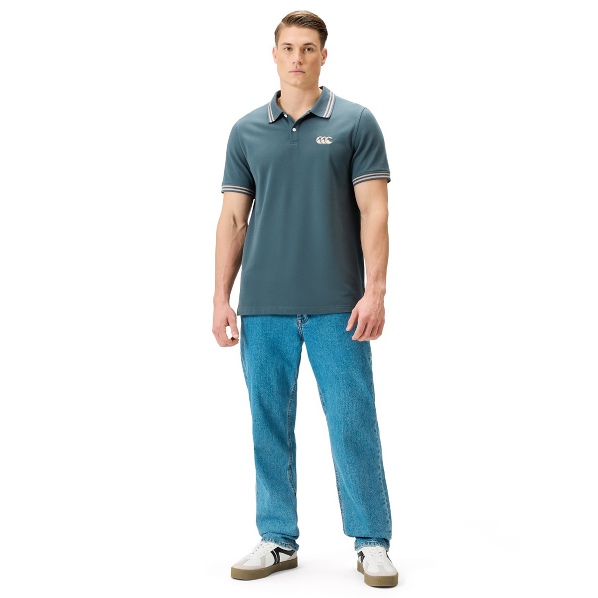 A man stands facing forward against a plain white background, wearing the Canterbury Solid Dye Polo by Canterbury in teal, paired with blue jeans and white sneakers.