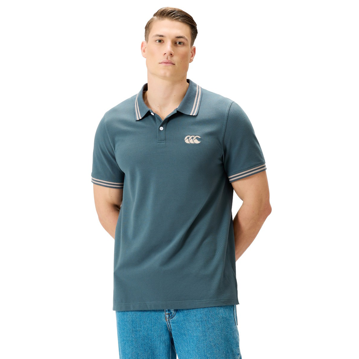 A person stands facing forward, wearing the Canterbury Solid Dye Polo by Canterbury—a teal short-sleeve polo featuring a striped collar and cuffs that evoke an athletic style—paired with light blue jeans, against a plain white background.