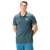 A person stands facing forward, wearing the Canterbury Solid Dye Polo by Canterbury—a teal short-sleeve polo featuring a striped collar and cuffs that evoke an athletic style—paired with light blue jeans, against a plain white background.