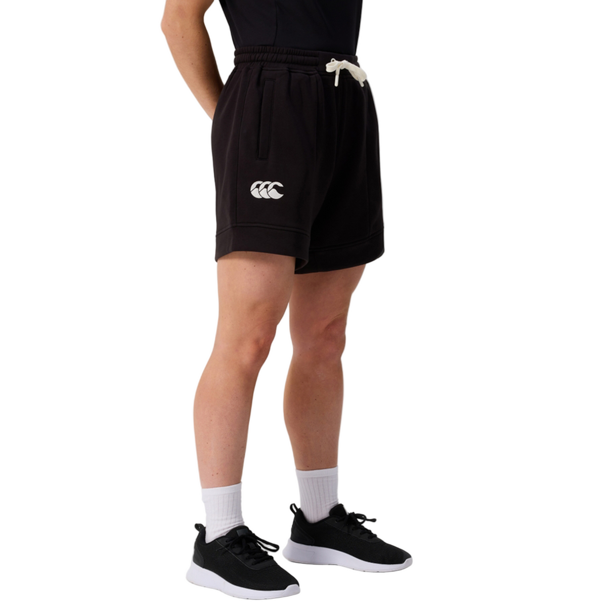 A person wears Canterbury Women's Ireland 25/26 Knit Shorts with a white logo, white socks, black sneakers, and a black top, standing against a plain light background.