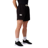 A person wears Canterbury Women's Ireland 25/26 Knit Shorts with a white logo, white socks, black sneakers, and a black top, standing against a plain light background.
