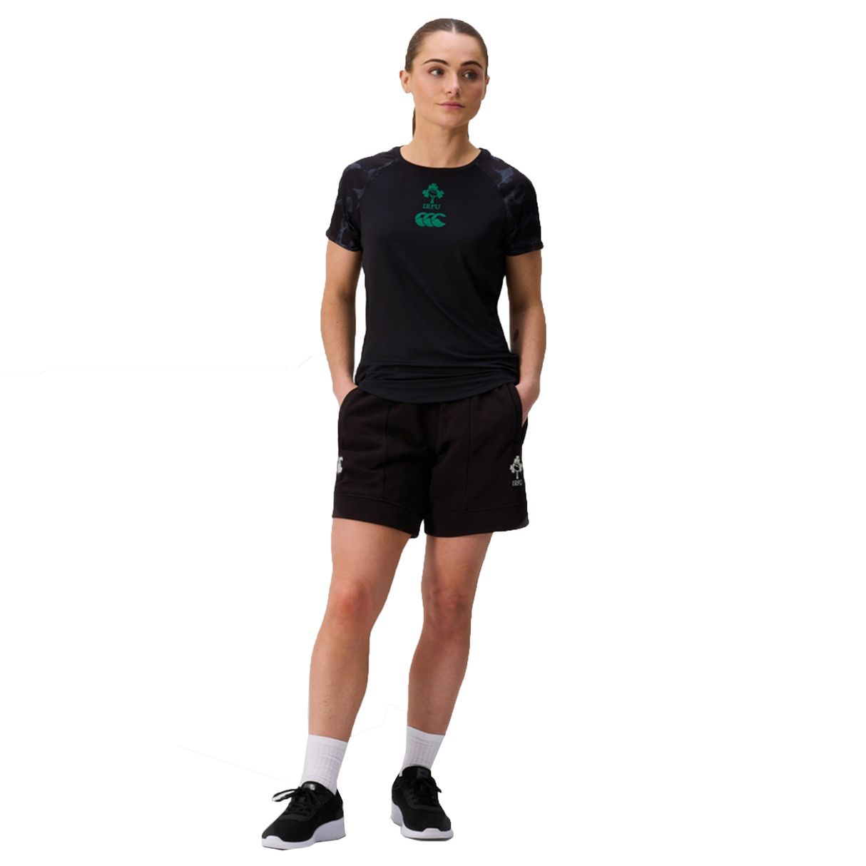 A person stands with hands in pockets, wearing a black athletic t-shirt, Canterbury Women's Ireland 25/26 Knit Short, white socks, and black sneakers against a white background.