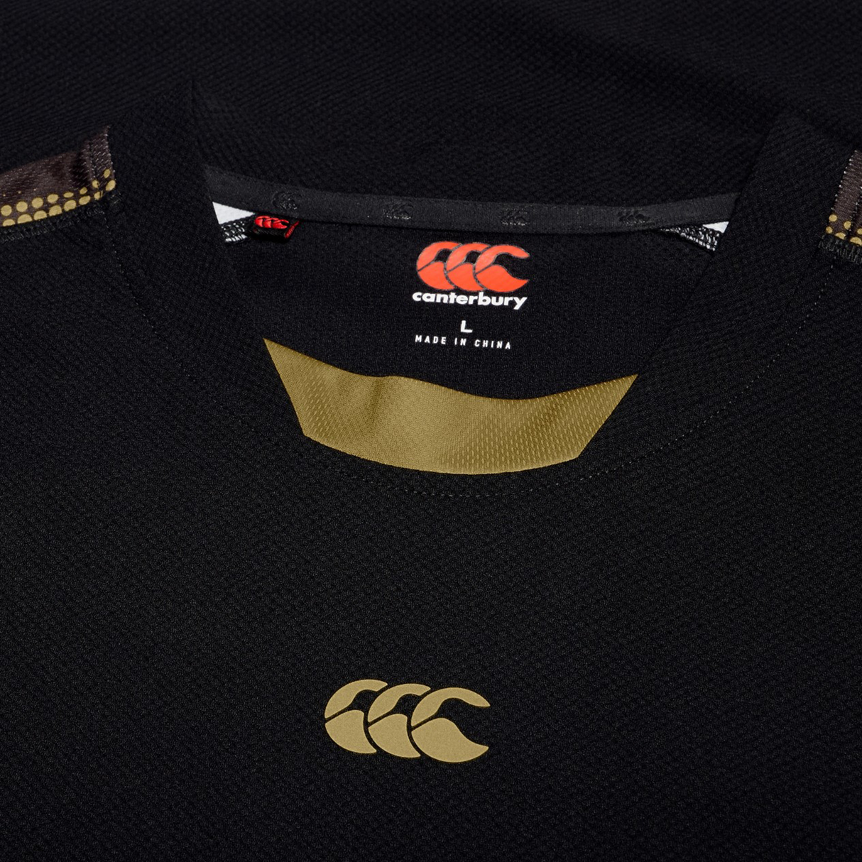 Close-up of a black Tempo+ Drill Top by Canterbury with gold logo and trim, neckline visible, and label showing size L and "Made in China.