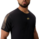 A man wears the Canterbury Tempo+ Drill Top, featuring a black design with a gold logo and patterned shoulder detail. Shown from the shoulders up against a white background. Made from moisture-wicking fabric for performance comfort.
